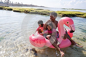 Father and two daughters ride flamingo buoy on the beach