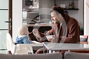 Father wearing bathrope spoon feeding hir infant baby boy child sitting in high chair at the dining table in kitchen at