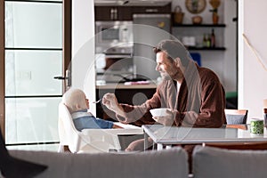 Father wearing bathrope spoon feeding hir infant baby boy child sitting in high chair at the dining table in kitchen at