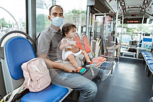 a father wearing a mask sits on a bench holding a cute little baby girl on the bus