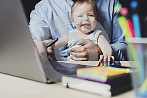 Father working on laptop with baby sitting on his knees.Toddler in front of laptop. Homebased online work for people with children