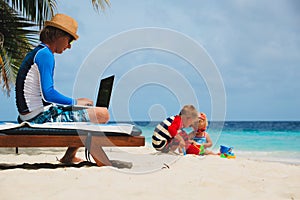 Father working on laptop while kids play at beach