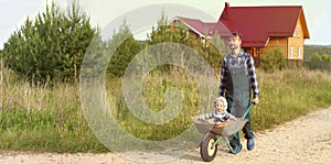 Father in working uniform pushing son in wheelbarrow on a rural road near the farm house. Family gardening concept. Banner, copy s