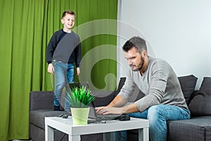 Father works on a laptop at home, his son bothers him. Businessman working from home and looking after a child, spending time with