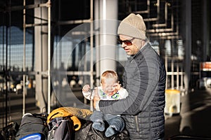 Fatherat comforting his crying infant baby boy child tired sitting on top of luggage cart in front of airport terminal