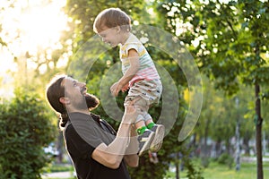 Fatherhood, parenthood, childhood, caring, summer and leisure concept - young dad with beard and long hair in black t