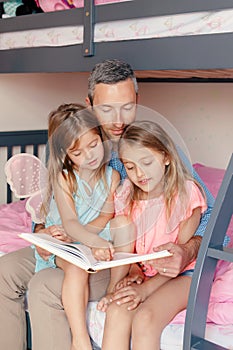 Fathers Day. Dad reading book to daughters girls. Family of three people sitting on a bed in bedroom. Happy father and children at
