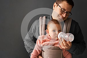 Fathre and his kid in baby carrier at the grey background wall, baby wearing concept