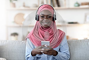 Favorite Leisure. Black muslim girl using smartphone and wireless headphones at home