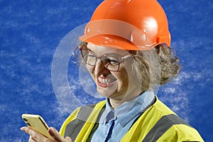 Feamle Engineer in the construction helmet with mobile phone on blue background, woman using smartphone app for warehouse