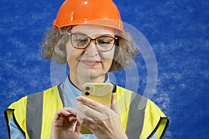 Feamle Engineer in the construction helmet and yellow vest with mobile phone and documents on blue background