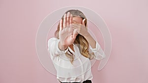 Fear-stricken, embarrassed young blonde woman hides her eyes, doing \'stop\' gesture in pink isolated background