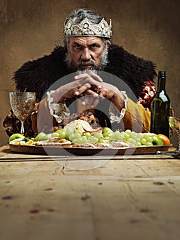 He feasts while the serfs starve. A mature king feasting alone in a banquet hall.