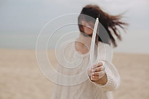 Feather in hand of beautiful woman with windy hair on background of sandy beach and sea, tranquil moment.  Free and wild. Stylish