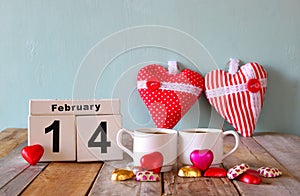 February 14th wooden vintage calendar with colorful heart shape chocolates next to couple cups on wooden table. selective focus