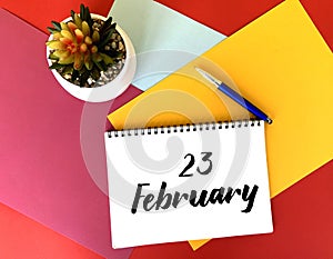 February 23 on a white notebook on a colorful bright background.Next to it is a potted flower and a blue pen.