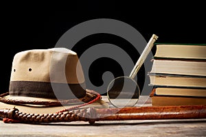 Fedora hat with bullwhip near magnifying glass and old books on black background. Adventure concept