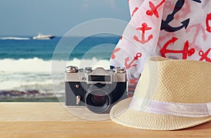 Fedora hat, old vintage camera and scarf over wooden table and sea landscape background. relaxation or vacation concept