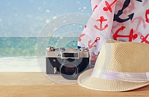 Fedora hat, old vintage camera and scarf over wooden table and sea landscape background. relaxation or vacation concept