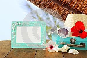 Fedora hat, sunglasses, tropical hibiscus flower next to blank frame over wooden table and beach landscape background