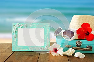 Fedora hat, sunglasses, tropical hibiscus flower next to blank frame over wooden table and beach landscape background