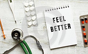 FEEL BETTER is written in a notebook on a white table next to pills and a stethoscope