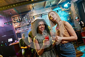 Feel the night here. Attractive young women looking at camera, posing with cocktail in their hands. Friends celebrating