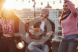 Feel the rhythm. Guitarist sits and singing for his friends at the rooftop with decorative colored light bulbs