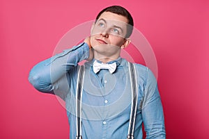 Portrait of attractive young man in blue shirt, suspenders and white bow tie touching his neck and looks pensive while standing