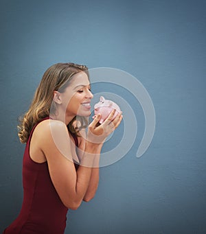 Feeling richer already. a young woman kissing a piggybank against a gray background.