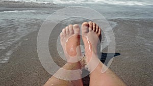 The feet of a child resting on a sandy beach against the backdrop of the sea. Waves with foam rolling up on the