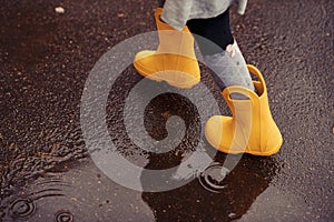 Feet of child in yellow rubber boots jumping over a puddle in th