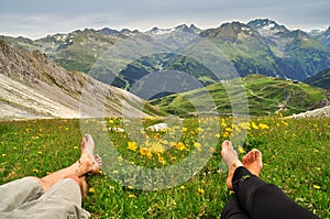 Feet from couple relaxing looking at snowy mountains surrounded by yellow flowers and green grass in Austria mountains