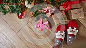 feet in funny Christmas socks Santa face wooden floor under fir tree gift boxes