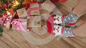 feet in funny Christmas socks Santa face wooden floor under fir tree gift boxes