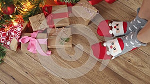 feet in funny Christmas socks Santa face wooden floor under fir tree gift boxes