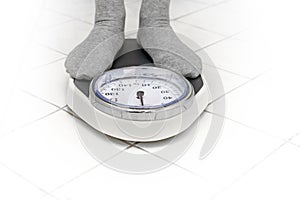 Feet in gray socks standing on a personal scale on the white tiled bathroom floor for weight measuring, copy space, selected focus