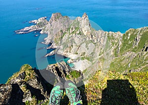 Feet in green sports shoes over the abyss, above the green rocky peninsula jutting out into the blue sea. Top view.