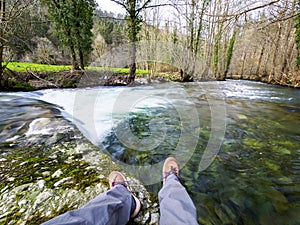 Feet and legs of an unrecognizable person sitting on a wall over a river with a beautiful landscape in the background and the