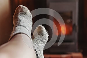 Feet legs in winter clothes wool socks at fireplace background. Woman sitting at home on winter or autumn evening relaxing and