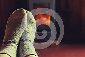 Feet legs in winter clothes wool socks at fireplace background. Woman sitting at home on winter or autumn evening relaxing and