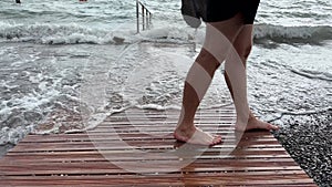 The feet of a man cross the frame against a wooden walkway going to the sea