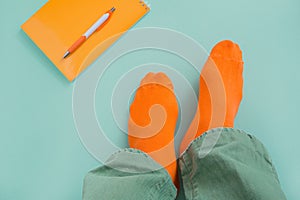 Feet of a man in orange socks and green jeans. Teal background with notepad and pen