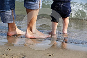 Feet of the men and baby standing in shallow water waiting for the wave