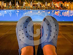 Feet in rubber blue flip flops, shoes in front of a pool of water in an all inclusive hotel at night in a warm tropical oriental