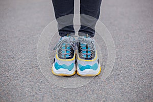Feet shod in sneakers multi-colored yellow, white, black and blue are on the asphalt road.