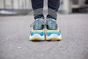 Feet shod in sneakers multi-colored yellow, white, black and blue are on the asphalt road.