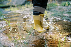Feet of a small child in yellow rubber boots stand in a stream sparkling in the sun. Cropped. Faceless