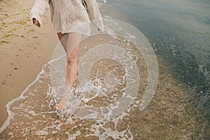 Feet in splashing waves. Carefree beautiful woman running barefoot on sandy beach with cold sea waves, having fun. Cropped view. S
