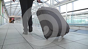 Feet of successful businessman walking in hall of terminal and pulling suitcase on wheels. Legs of young business person